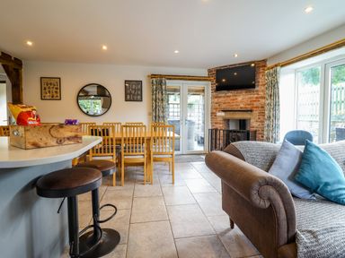An open living and dining area with a brown sofa and blue cushions a dining table with wooden chairs a brick fireplace with a mounted TV and large windows at Y Stabl (The Stable) in Llanrhaeadr-Ym-Mochnant