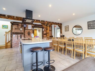 A kitchen island with two stools and a long dining table with chairs in a room with exposed brick and wooden beams at Y Stabl (The Stable) in Llanrhaeadr-Ym-Mochnant
