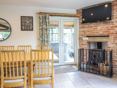 A dining area with wooden chairs and table near a set of glass doors and a brick fireplace with a mounted television at Y Stabl The Stable in Llanrhaeadr-Ym-Mochnant