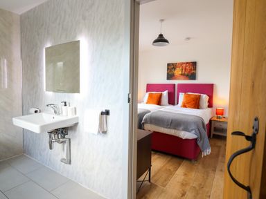 A bathroom with a sink and mirror next to a bedroom with two single beds with orange pillows at Y Stabl in Llanrhaeadr-Ym-Mochnant