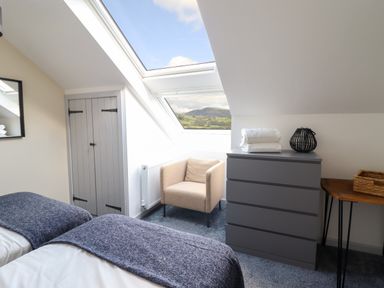 A bedroom with two beds a beige armchair a gray chest of drawers and a small wooden table at Y Stabl The Stable in Llanrhaeadr-Ym-Mochnant