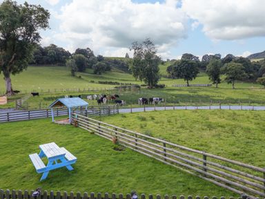 A fenced grassy area with a blue picnic table and covered sandbox with cows grazing in fields beyond at Y Stabl (The Stable) in Llanrhaeadr-Ym-Mochnant