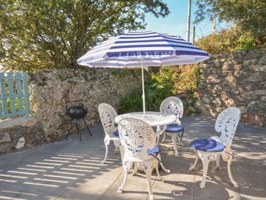 An outdoor patio area with a white metal table and four chairs with blue cushions and a blue and white striped umbrella and a small black barbecue grill by a stone wall at Ffrwd in Rhosneigr