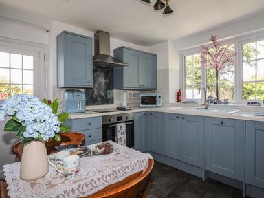 A kitchen with blue cabinets a table with flowers and mugs and a window with a pink flower decoration at Ffrwd in Rhosneigr