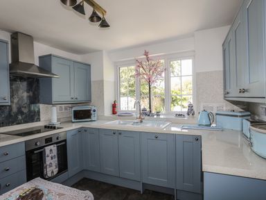 A kitchen with blue cabinets white countertops a window above the sink and various appliances at Ffrwd in Rhosneigr