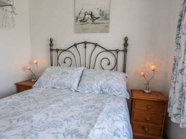 A bedroom with a metal bed frame, two wooden bedside tables with lamps, floral bedding, and wall art at Ffrwd in Rhosneigr
