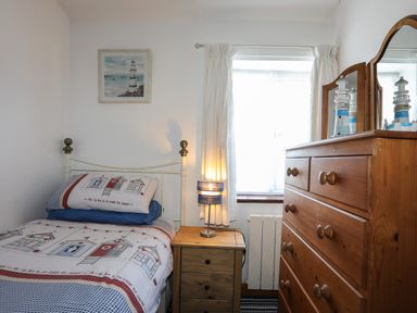A bedroom with a single bed with seaside themed bedding a wooden nightstand with a lamp a window with white curtains a wooden dresser with a mirror and a lighthouse ornament at Ffrwd in Rhosneigr