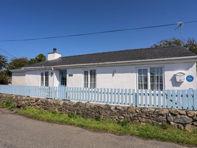 A white single-story house with a blue door and blue picket fence on a stone wall by a road at Ffrwd in Rhosneigr