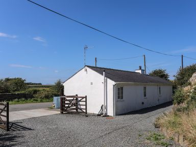 A white single-story house with a gray roof and wooden gates on a gravel driveway at Ffrwd in Rhosneigr