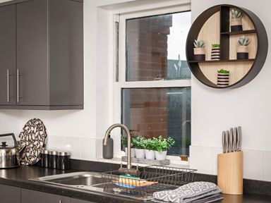 A kitchen sink with a drying rack cutting knives in a wooden block and a wall shelf with small plants at Sunnymeade in Buxton Norfolk