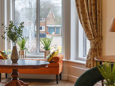A seating area with an orange bench sofa cushions and plants near a window at Sunnymeade in Buxton Norfolk