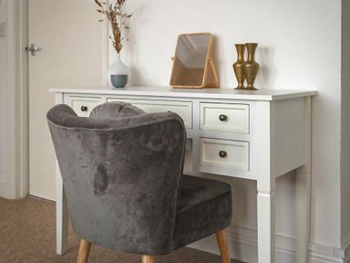 A grey upholstered chair in front of a white desk with a small mirror a vase and two metal vases on top at Sunnymeade in Buxton Norfolk