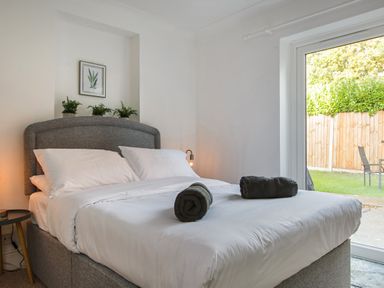 A bedroom with a grey upholstered bed white bedding two rolled towels green plants and a sliding glass door at Sunnymeade in Buxton Norfolk