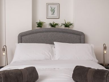 A bedroom with a bed with white pillows and sheets two brown towels on the bed three potted plants on the headboard two bedside lamps and a framed picture on the wall at Sunnymeade in Buxton Norfolk