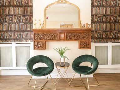 Two green chairs and a small round table with a plant in front of a decorative wooden mantle and mirror in a room with bookshelves wallpaper at Sunnymeade in Buxton Norfolk