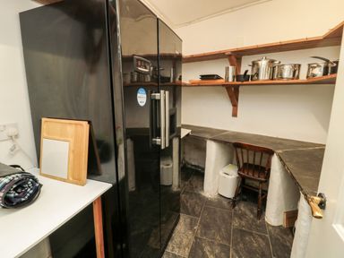 A small kitchen area with a black refrigerator wooden shelves with pots and pans a wooden chair and a white countertop at Jubilee House in Embleton