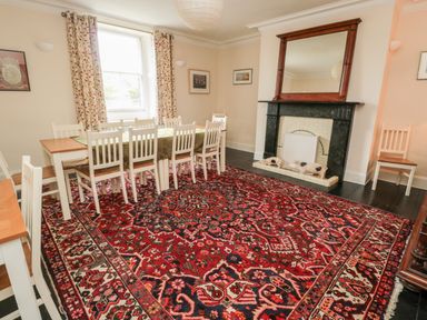 A dining room with a long table and white chairs a fireplace with a mirror above and pig figurines a large patterned rug on the floor at Jubilee House in Embleton