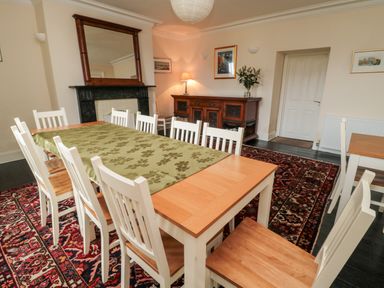 A dining room with a long wooden table covered by a green table runner surrounded by white chairs a wooden sideboard with a lamp and flowers a fireplace with a mirror above and a patterned rug on the floor at Jubilee House in Embleton