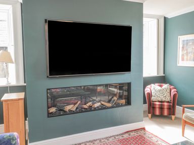 A living room with a wall-mounted television above a fireplace a striped armchair with a patterned cushion and a wooden side table near windows at Jubilee House in Embleton