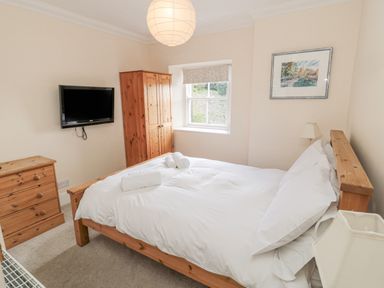 A bedroom with a wooden bed and white bedding a wooden dresser and wardrobe a wall mounted tv and a window with blinds at Jubilee House in Embleton