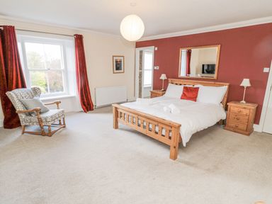 A bedroom with a wooden bed and two bedside tables with lamps a patterned armchair and a window with red curtains at Jubilee House in Embleton