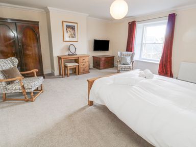A bedroom with a bed with white linens two armchairs a wardrobe a dressing table a chest a wall-mounted TV and a window with red curtains at Jubilee House in Embleton