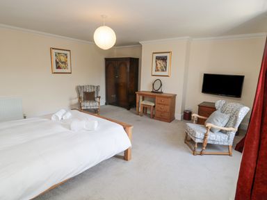 A bedroom with a bed with white linens two patterned armchairs a wooden wardrobe a wooden dressing table and a wall-mounted TV at Jubilee House in Embleton