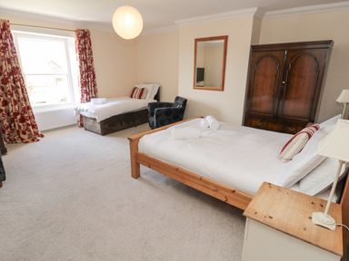 A bedroom with two beds a wooden wardrobe an armchair and a window with red floral curtains at Jubilee House in Embleton