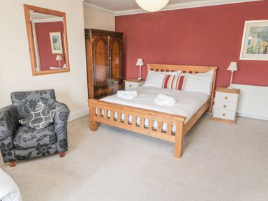A bedroom with a wooden bed with white linens two nightstands with lamps a wooden wardrobe a grey armchair and a wall mirror at Jubilee House in Embleton