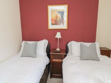 A bedroom with two single beds with white bedding and patterned pillows a wooden nightstand with a lamp and a remote between the beds and a framed picture on a red wall at Jubilee House in Embleton