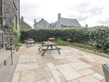 A patio with a wooden picnic table chairs stone paving and a hedge surrounding the area at Jubilee House in Embleton