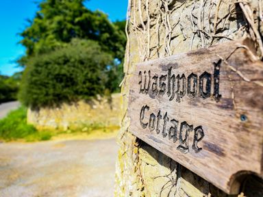 A wooden sign reading Washpool Cottage on a stone wall with greenery and a road in the background at Washpool in Milton-Under-Wychwood