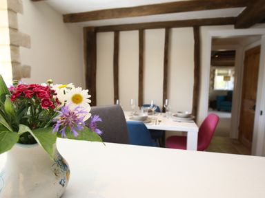 A dining room with a white table set with plates and glasses and colorful chairs and a vase of flowers on a counter at Washpool in Milton-Under-Wychwood