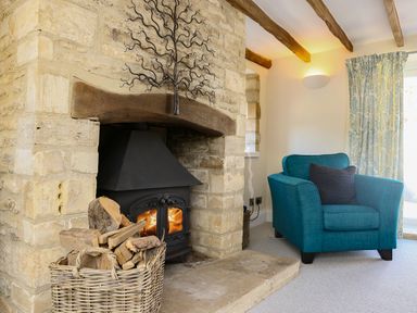 A stone fireplace with a wood-burning stove a basket of firewood a blue armchair and floral curtains at Washpool Milton-Under-Wychwood