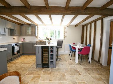 A kitchen with grey cabinets an island with wine storage a dining table with colorful chairs and exposed wooden beams at Washpool in Milton-Under-Wychwood