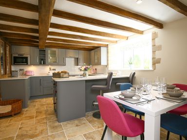 A kitchen with gray cabinets wooden ceiling beams a white dining table with colorful chairs and tableware at Washpool in Milton-Under-Wychwood