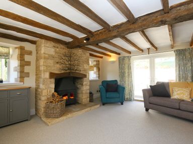 A living room with a stone fireplace a blue armchair a gray sofa with cushions and wooden ceiling beams at Washpool in Milton-Under-Wychwood