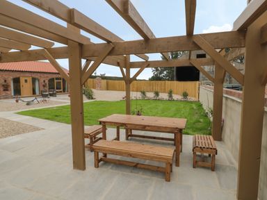 An outdoor seating area with wooden benches and a table under a wooden pergola with a garden and brick building in the background at Plough Cottage in Little Kelk near Bridlington