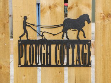 A black metal sign with a farmer plowing with a horse saying Plough Cottage on a wooden fence at Plough Cottage in Little Kelk near Bridlington