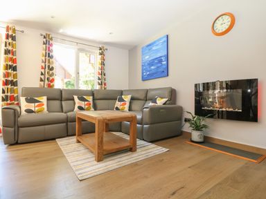 A living room with a grey sectional sofa and patterned cushions a wooden coffee table striped rug a wall clock a potted plant a blue painting and a wall-mounted fireplace at 32 Trevithick in Hayle