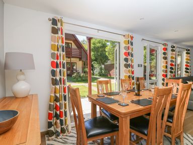 A dining area with wooden table and chairs set with placemats and glasses near patterned curtains and large glass doors opening to a garden at 32 Trevithick in Hayle