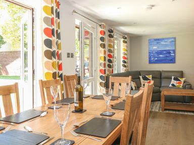 A dining room with a wooden table set for six and a seating area with a grey sofa and colorful curtains at 32 Trevithick in Hayle