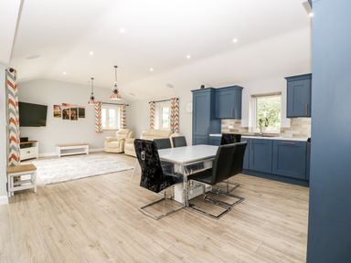 An open-plan room with blue kitchen cabinets a dining table with black chairs beige sofas a wall-mounted TV and patterned curtains at Strawberry Stables in Iwood near Congresbury