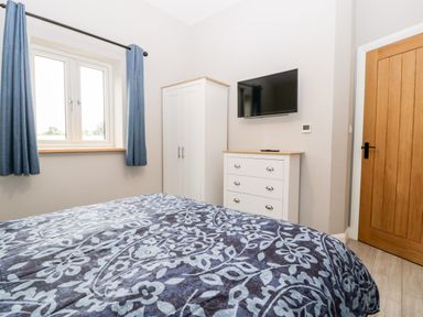 A bedroom with a bed blue patterned blanket a white wardrobe white chest of drawers a wall-mounted tv and a wooden door at Strawberry Stables in Iwood near Congresbury