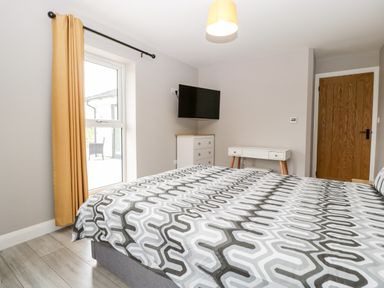 A bedroom with a patterned bedspread a window with yellow curtains a wall-mounted tv a wooden door and white furniture at Strawberry Stables in Iwood near Congresbury