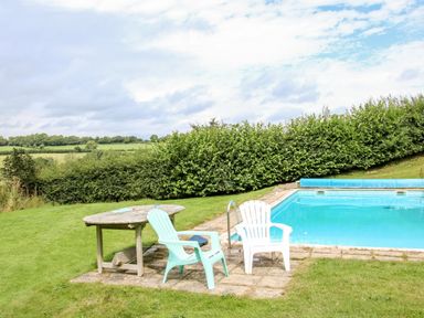 An outdoor area with a swimming pool two chairs and a wooden table overlooking green fields at Callow Fold in Dinchope near Craven Arms