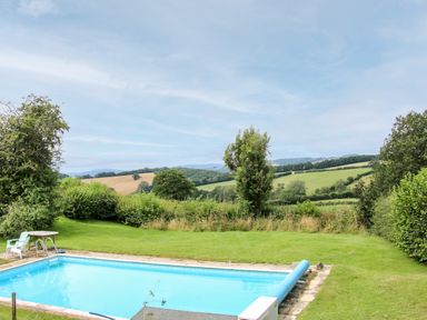 An outdoor swimming pool with a chair and table beside it and green fields and trees in the background at Callow Fold in Dinchope near Craven Arms
