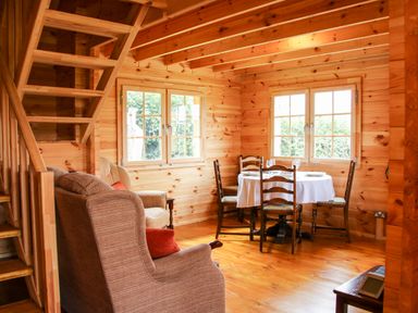 A wooden dining area with a round table covered by a white tablecloth and four chairs next to two windows in a log cabin