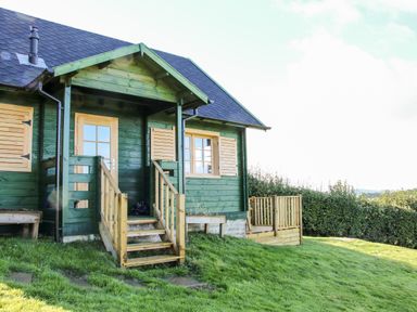 A green wooden cabin with wooden door and windows shutters on a grassy area at Callow Fold in Dinchope near Craven Arms