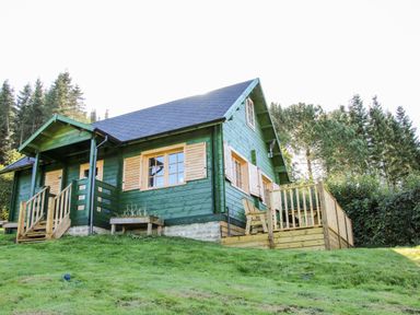 A green wooden cabin with wooden shutters and a deck surrounded by trees at Callow Fold in Dinchope near Craven Arms